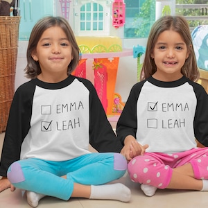 Peut inclure: Deux enfants portant des chemises de baseball blanches et noires avec le texte "Emma" et "Leah" dessus. Une chemise a une coche &agrave; c&ocirc;t&eacute; de "Emma" et l'autre a une coche &agrave; c&ocirc;t&eacute; de "Leah".