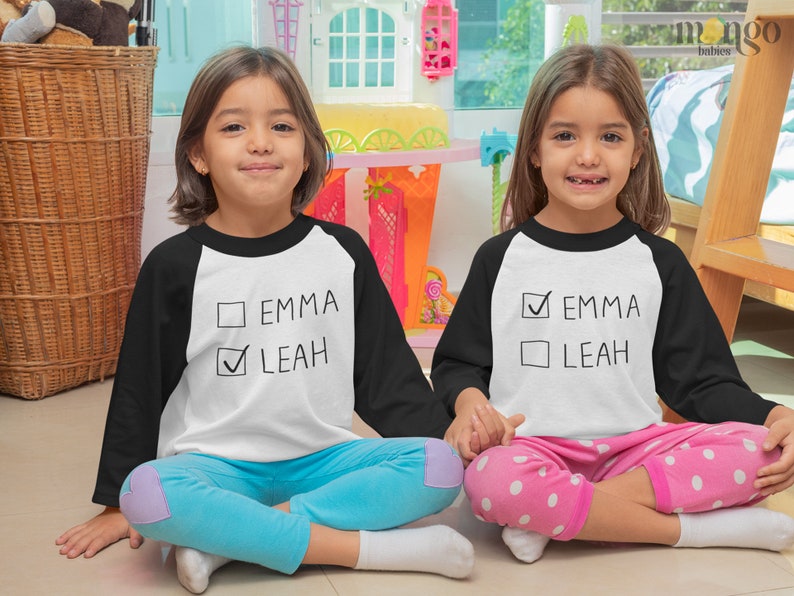 Peut inclure: Deux enfants portant des chemises de baseball blanches et noires avec le texte "Emma" et "Leah" dessus. Une chemise a une coche &agrave; c&ocirc;t&eacute; de "Emma" et l'autre a une coche &agrave; c&ocirc;t&eacute; de "Leah".