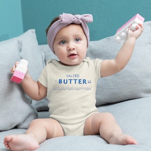 May include: A baby wearing a cream-colored onesie with the words "SALTED BUTTER" printed in blue. The baby is holding pink and white toy blocks and wearing a pink headband.
