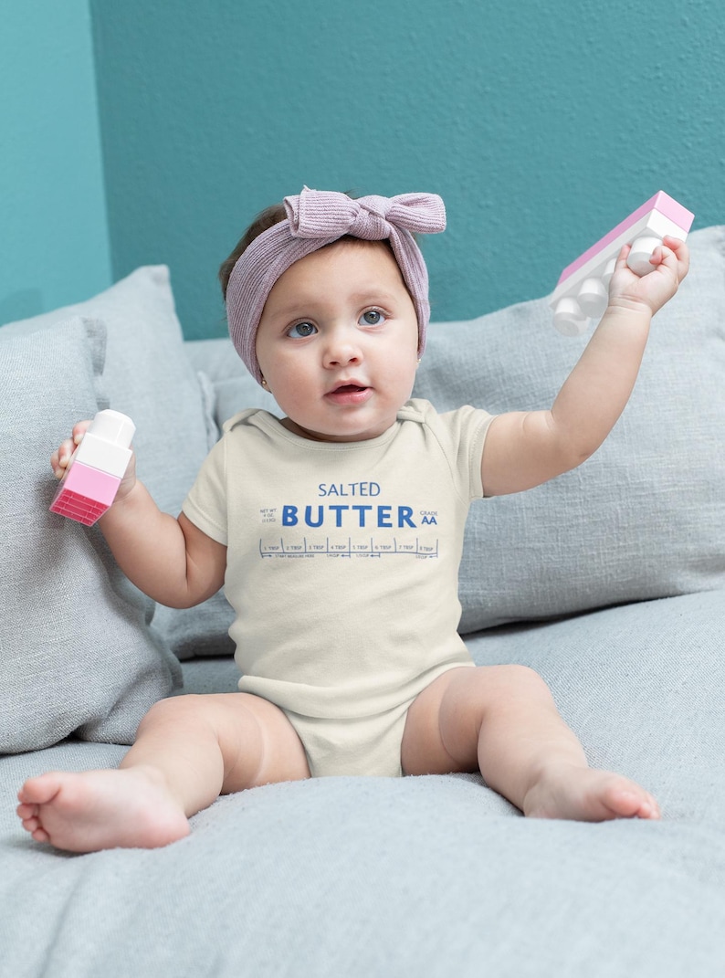 May include: A baby wearing a cream-colored onesie with the words "SALTED BUTTER" printed in blue. The baby is holding pink and white toy blocks and wearing a pink headband.