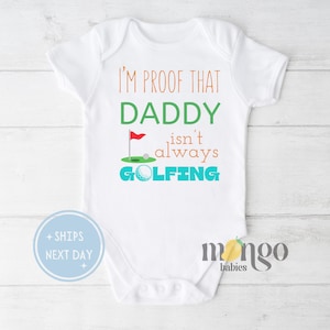 May include: White baby onesie with the text "I'm proof that Daddy isn't always golfing" in orange and blue. A golf ball and flag are illustrated on the onesie.