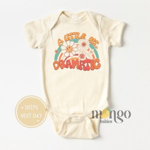 May include: A cream-colored baby onesie with a retro-style graphic of a rainbow with flowers and the text "A little bit dramatic".