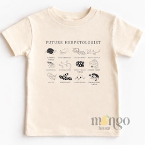 May include: A cream-coloured short-sleeve t-shirt featuring a black graphic print with the text FUTURE HERPETOLOGIST. The design includes a grid of twelve illustrations of reptiles and amphibians with their corresponding names printed underneath each.