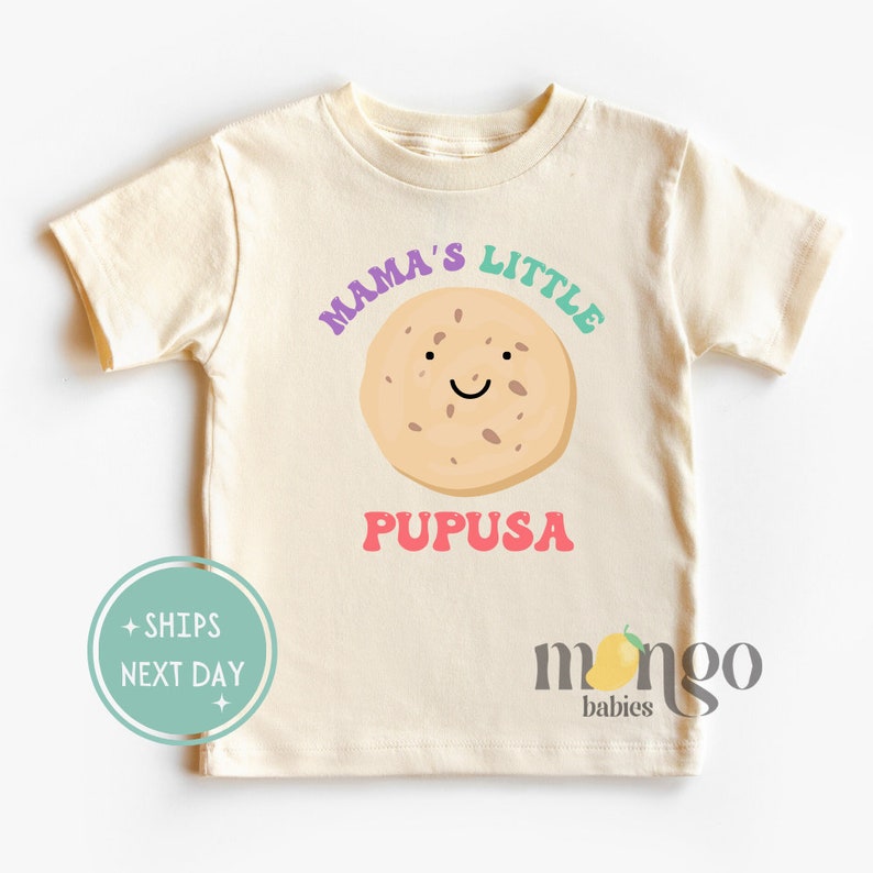 K&ouml;nnte beinhalten: Ein hellbeiges T-Shirt mit kurzen &Auml;rmeln mit dem Text "Mama's Little Pupusa" in bunten Buchstaben. Ein Cartoon-Pupusa mit einem l&auml;chelnden Gesicht befindet sich in der Mitte des Textes. Der Text "mango babies" befindet sich in der rechten unteren Ecke des Shirts.