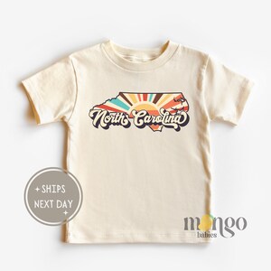 Puede incluir: Una camiseta de manga corta de color crema con un gr&aacute;fico retro del estado de Carolina del Norte con un dise&ntilde;o de rayos de sol y el texto "North Carolina".