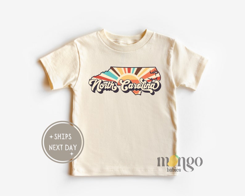 Puede incluir: Una camiseta de manga corta de color crema con un gr&aacute;fico retro del estado de Carolina del Norte con un dise&ntilde;o de rayos de sol y el texto "North Carolina".