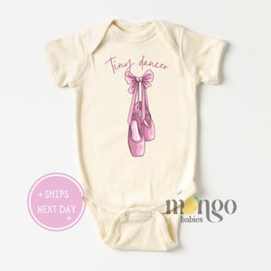 May include: A cream colored baby onesie with short sleeves and the text "Tiny dancer" printed on the front. A pink bow with pink ballet shoes hanging from it are also printed on the onesie.