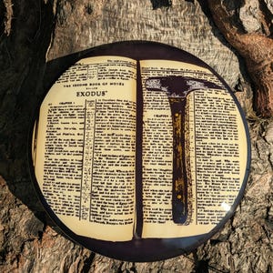 May include: A circular, black and tan decorative plate with an open book design, text from Exodus, and a hammer illustration. The plate is set against a natural wood background.
