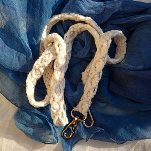 Könnte beinhalten: Ein weißes gehäkeltes Band mit goldenen Metallschließen. Das Band liegt auf einem blauen Stoffhintergrund.