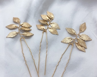 May include: Three gold-colored hair pins, each featuring a cluster of textured leaves. The leaves are attached to a wire stem, which is secured to a long, wavy pin. The pins are arranged on a white, textured surface.