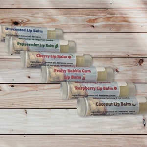 May include: Six lip balm tubes are arranged diagonally on a wooden surface. Each tube has a label with a different flavour: Unscented, Peppermint, Cherry, Fruity Bubble Gum, Raspberry, and Coconut. The labels also list ingredients.