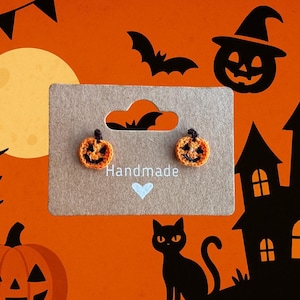 May include: Handmade Halloween-themed earrings. The earrings are shaped like small orange pumpkins with black facial features. They are displayed on a brown card with the word "Handmade" and a blue heart. The background features Halloween decorations.