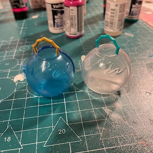 May include: Two clear, spherical ornaments with handles. One is transparent, the other is blue. Both have a wave design. Paint bottles are in the background.