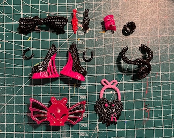 May include: A selection of miniature, handcrafted accessories in black and pink, including a bow, high-heeled shoes, a bat-shaped ring, and a heart-shaped lock. The items are displayed on a green cutting mat.