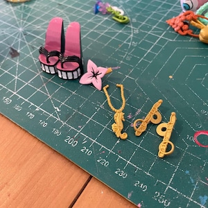 Könnte beinhalten: Pinke Plateausandalen mit schwarzen Riemen und weiß-schwarzen Sohlen. Eine rosa Blume und eine goldene Seepferdchen-Anhänger-Halskette sind ebenfalls zu sehen, zusammen mit zwei goldenen Schlüsselanhängern. Auf einer grünen Schneidematte.