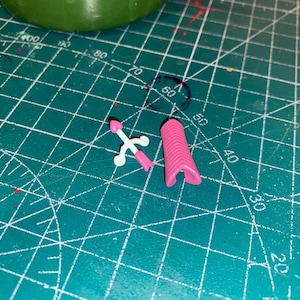 May include: A small pink and white toy sword and a pink, ridged object are on a green cutting mat. The sword has a white handle and a pink blade. The other object is a pink, curved shape.