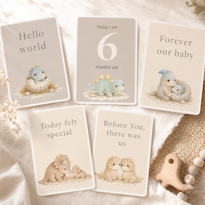 May include: Set of five baby milestone cards with dinosaur illustrations. Cards include text: "Hello world," "Today I am 6 months old," "Forever our baby," "Today felt special," and "Before You, there was us." The cards have a neutral color palette.