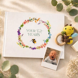 May include: A white baby memory book with the text "YOUR FIRST YEARS" surrounded by a floral wreath and a bee illustration. A crochet bee rattle and a photo of a baby are next to the book.