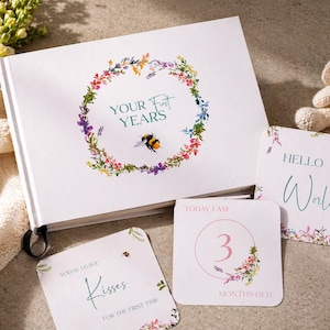 May include: A white baby memory book with the text "YOUR FIRST YEARS" surrounded by a floral wreath and a bumblebee. Includes milestone cards with floral accents and the text "HELLO World", "3 MONTHS OLD", and "TODAY I GAVE Kisses". A stuffed bunny is also present.