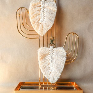 Pode incluir: Dois chaveiros de pena de macramé branco pendurados em um suporte de metal dourado em forma de cacto. O suporte está sobre uma superfície marrom.