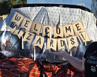 Może przedstawiać: Brązowo-biały baner z napisem "WELCOME WIZARDS!" wiszący na samochodzie. Baner jest wykonany z papieru i ma vintage'owy wygląd. Samochód jest ozdobiony w stylu Harry'ego Pottera.