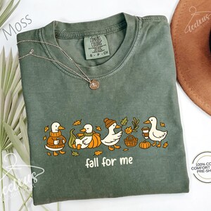 a green t-shirt with a graphic design of ducks and fall-themed elements, such as leaves and pumpkins, along with the text "fall for me". The t-shirt is laid out on a white surface, with a brown hat and some greenery visible in the background.