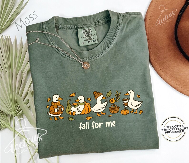 a green t-shirt with a graphic design of ducks and fall-themed elements, such as leaves and pumpkins, along with the text "fall for me". The t-shirt is laid out on a white surface, with a brown hat and some greenery visible in the background.