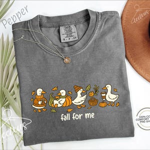 a gray t-shirt with a graphic design of ducks wearing hats and holding various objects, such as a pumpkin and a basket, along with the text "fall for me". The t-shirt is laid out on a white surface, with a brown hat and some leaves in the background.
