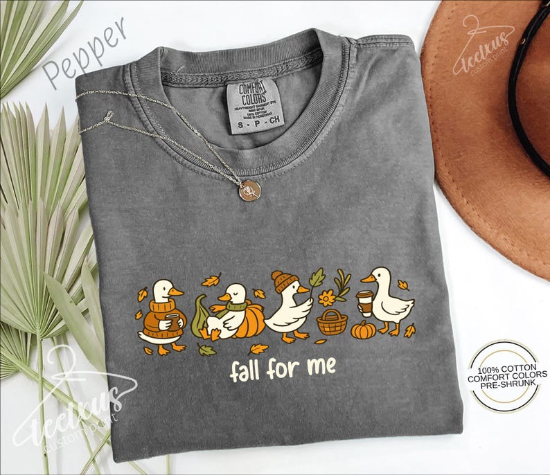 a gray t-shirt with a graphic design of ducks wearing hats and holding various objects, such as a pumpkin and a basket, along with the text "fall for me". The t-shirt is laid out on a white surface, with a brown hat and some leaves in the background.