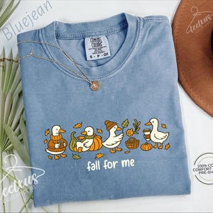 A blue t-shirt with a graphic design of ducks wearing fall-themed outfits and holding autumn-colored objects, accompanied by the text "fall for me". The t-shirt is displayed on a white background, with a brown hat and some greenery visible in the background.