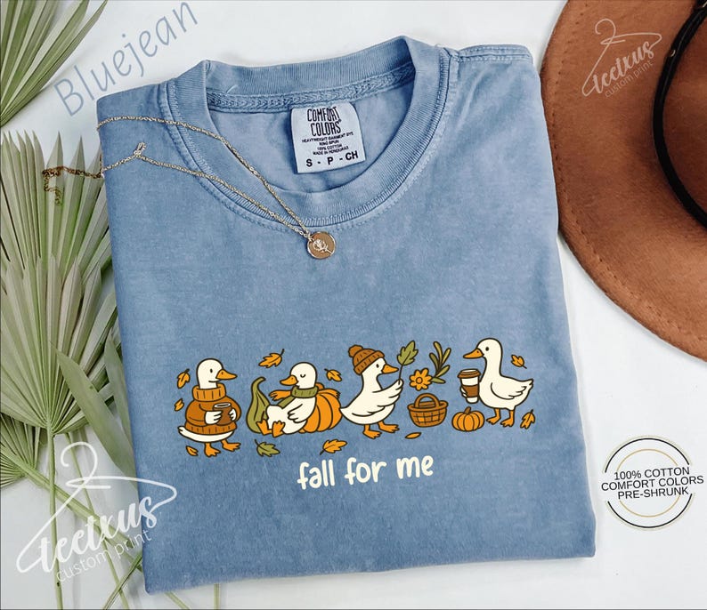 A blue t-shirt with a graphic design of ducks wearing fall-themed outfits and holding autumn-colored objects, accompanied by the text "fall for me". The t-shirt is displayed on a white background, with a brown hat and some greenery visible in the background.