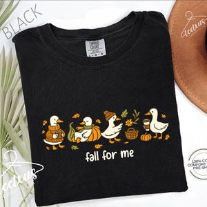 a black t-shirt with a graphic design of ducks wearing autumn-themed clothing and accessories, including a pumpkin and a basket. The phrase "fall for me" is written below the design.