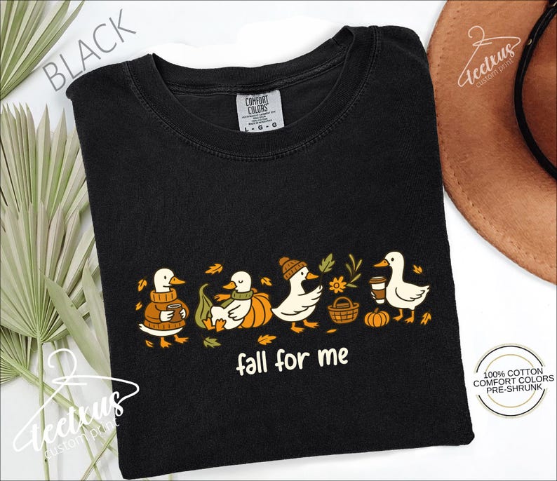 a black t-shirt with a graphic design of ducks wearing autumn-themed clothing and accessories, including a pumpkin and a basket. The phrase "fall for me" is written below the design.
