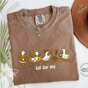 a brown t-shirt with a graphic design of ducks and fall-themed elements, such as pumpkins and leaves, along with the text "fall for me". The t-shirt is laid out on a white surface, with a brown hat and some greenery visible in the background.