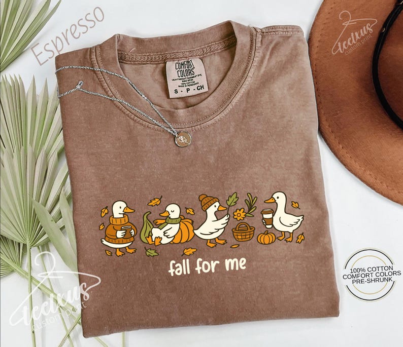 a brown t-shirt with a graphic design of ducks and fall-themed elements, such as pumpkins and leaves, along with the text "fall for me". The t-shirt is laid out on a white surface, with a brown hat and some greenery visible in the background.