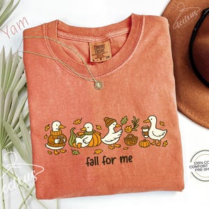 a pink t-shirt with a graphic design of ducks and fall-themed elements, such as pumpkins and leaves, along with the text "fall for me". The t-shirt is displayed on a white background, with a brown hat and some greenery visible in the background.