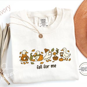 a white t-shirt with a graphic design of ducks wearing fall-themed outfits, accompanied by the text "fall for me". The t-shirt is displayed alongside other fall-themed items, such as a hat and some dried leaves.