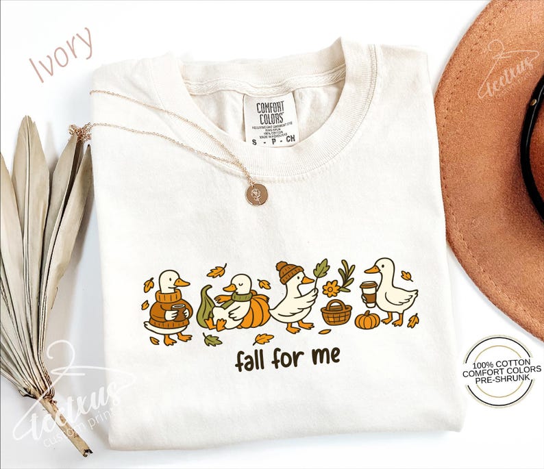a white t-shirt with a graphic design of ducks wearing fall-themed outfits, accompanied by the text "fall for me". The t-shirt is displayed alongside other fall-themed items, such as a hat and some dried leaves.