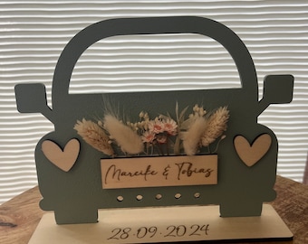 May include: A light blue wooden car cutout with a white base. The car has two white hearts on the sides and a sign that says "Marcelle & Tobias" with dried flowers in the back. The base has the date "28.09.2024" written on it.