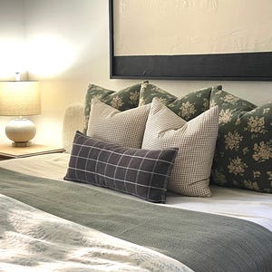 K&ouml;nnte beinhalten: Ein Bett mit wei&szlig;en Bettw&auml;sche und einer gr&uuml;nen Tagesdecke. Auf dem Bett liegen mehrere Kissen, darunter ein schwarzes und wei&szlig;es Kissen mit Muster, ein braunes und wei&szlig;es Karo-Kissen und ein gr&uuml;nes Blumenkissen.