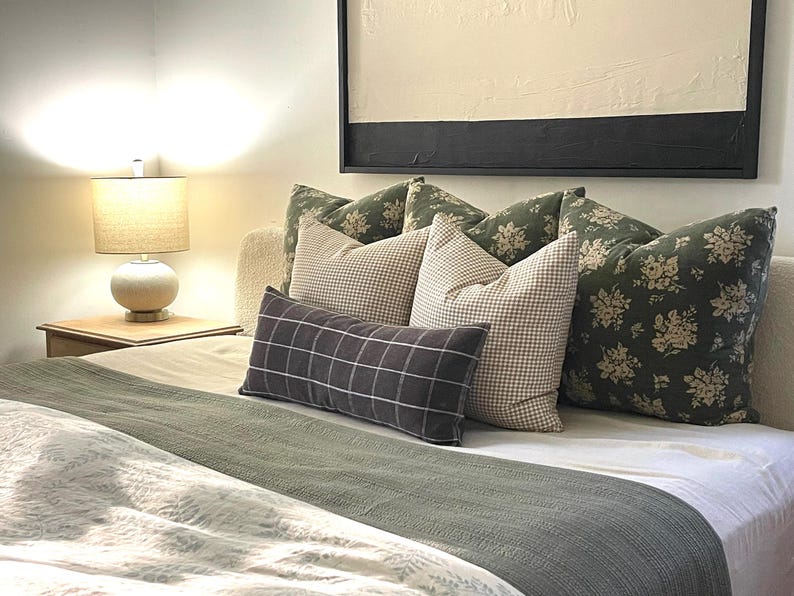 K&ouml;nnte beinhalten: Ein Bett mit wei&szlig;en Bettw&auml;sche und einer gr&uuml;nen Tagesdecke. Auf dem Bett liegen mehrere Kissen, darunter ein schwarzes und wei&szlig;es Kissen mit Muster, ein braunes und wei&szlig;es Karo-Kissen und ein gr&uuml;nes Blumenkissen.
