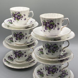 Peut inclure: Une pile de six tasses à thé et soucoupes avec un motif floral violet. Les tasses et les soucoupes sont blanches avec une bordure dorée.