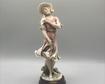 Könnte beinhalten: Eine Porzellanfigur einer Frau in einem rosa Kleid und einem weißen Hut, die eine weiße Taube hält. Sie steht auf einem Sockel mit grünem Laub und Blumen. Der Sockel ist aus Holz und trägt ein Etikett mit der Aufschrift "Lladro".