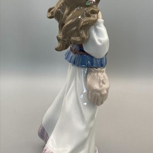 Puede incluir: Figurilla de porcelana de una ni&ntilde;a con un vestido blanco con estampado floral, un top azul y blanco y una corona de flores. Lleva una peque&ntilde;a bolsa en la mano.