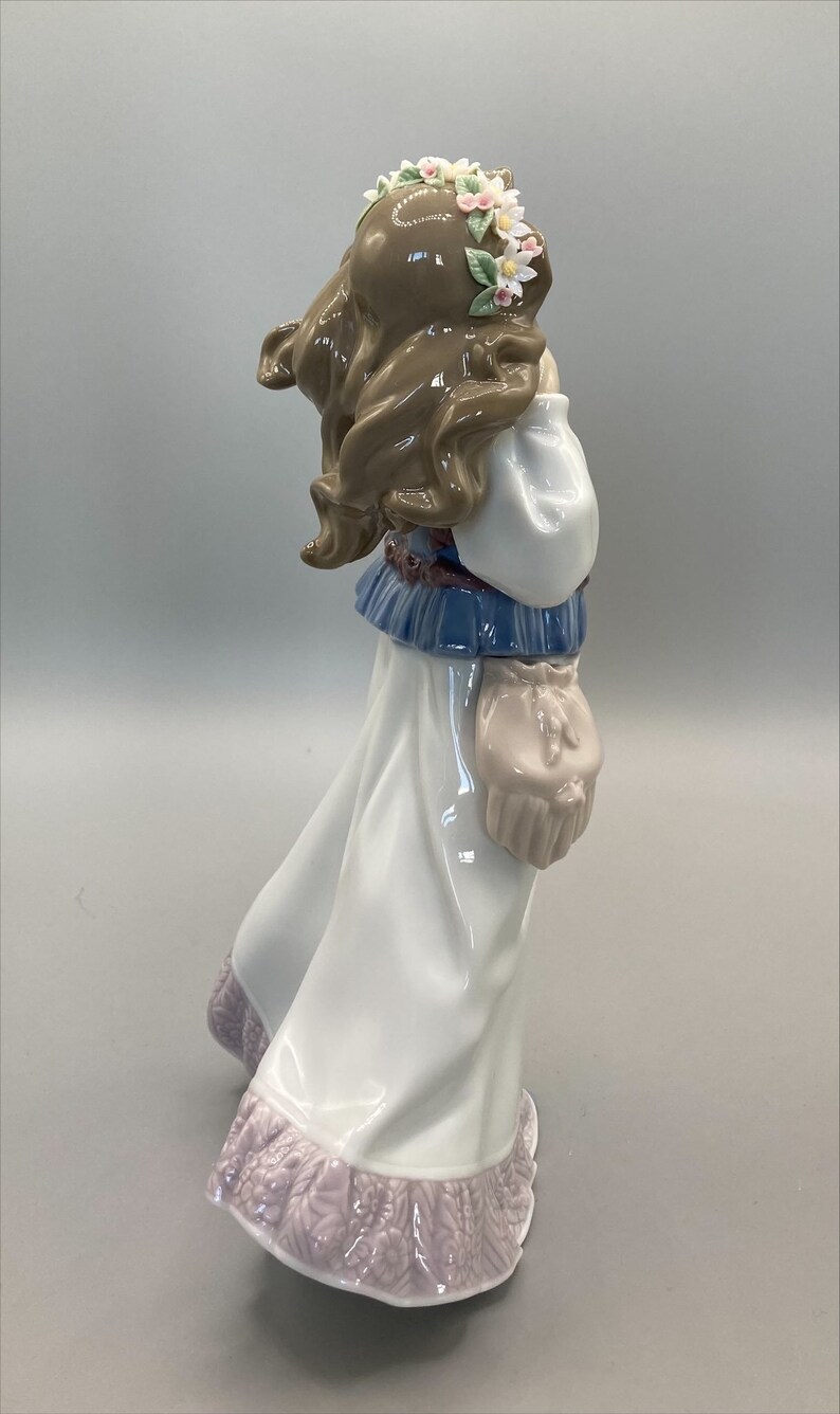 Puede incluir: Figurilla de porcelana de una ni&ntilde;a con un vestido blanco con estampado floral, un top azul y blanco y una corona de flores. Lleva una peque&ntilde;a bolsa en la mano.