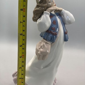 Puede incluir: Una figurita de porcelana de una ni&ntilde;a que lleva un vestido blanco con un chaleco azul y una corona de flores. Lleva una cesta en la mano y tiene una dulce expresi&oacute;n en su rostro.