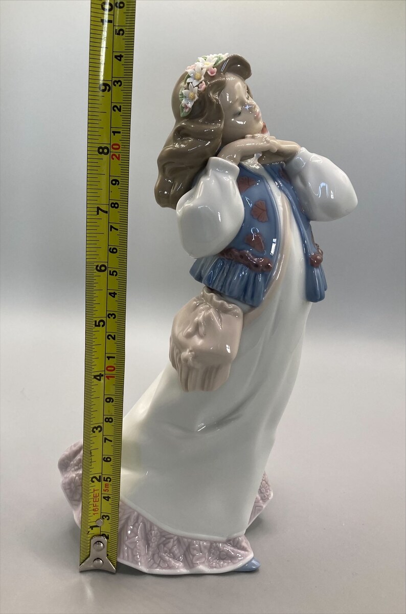 Puede incluir: Una figurita de porcelana de una ni&ntilde;a que lleva un vestido blanco con un chaleco azul y una corona de flores. Lleva una cesta en la mano y tiene una dulce expresi&oacute;n en su rostro.