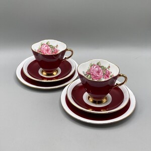 Può includere: Due tazzine da tè e piattini con un motivo floreale. Le tazzine e i piattini sono di un rosso bordeaux intenso con un bordo bianco e dettagli dorati. Il motivo floreale presenta rose rosa.