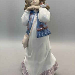Puede incluir: Una figurita de porcelana de una ni&ntilde;a con cabello casta&ntilde;o que lleva un vestido blanco con un chaleco azul y una corona de flores rosas. Ella sostiene un peque&ntilde;o p&aacute;jaro en su mano.