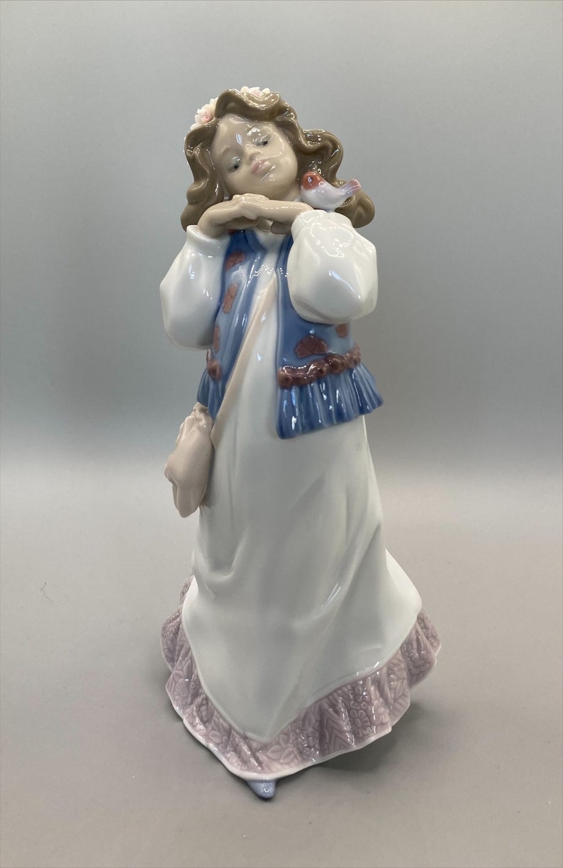 Puede incluir: Una figurita de porcelana de una ni&ntilde;a con cabello casta&ntilde;o que lleva un vestido blanco con un chaleco azul y una corona de flores rosas. Ella sostiene un peque&ntilde;o p&aacute;jaro en su mano.
